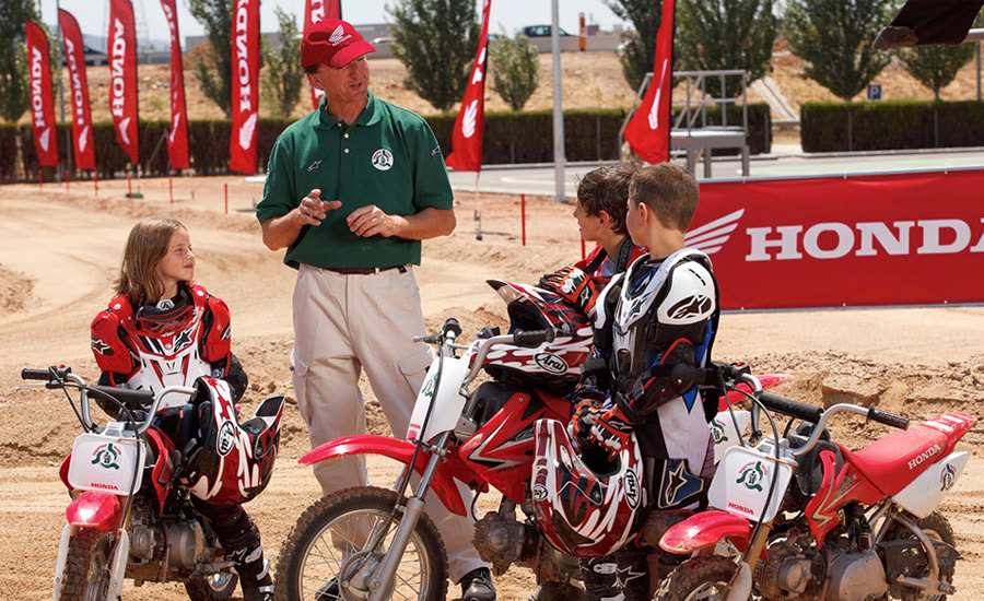 Tres niños con equipo Honda escuchan a un profesor del curso de conducción infantil encima de sus motos Honda