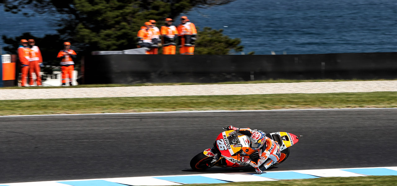 Dani Pedros tomando una curva de lado en Phillip Island, detrás operarios de pista observan frente al mar