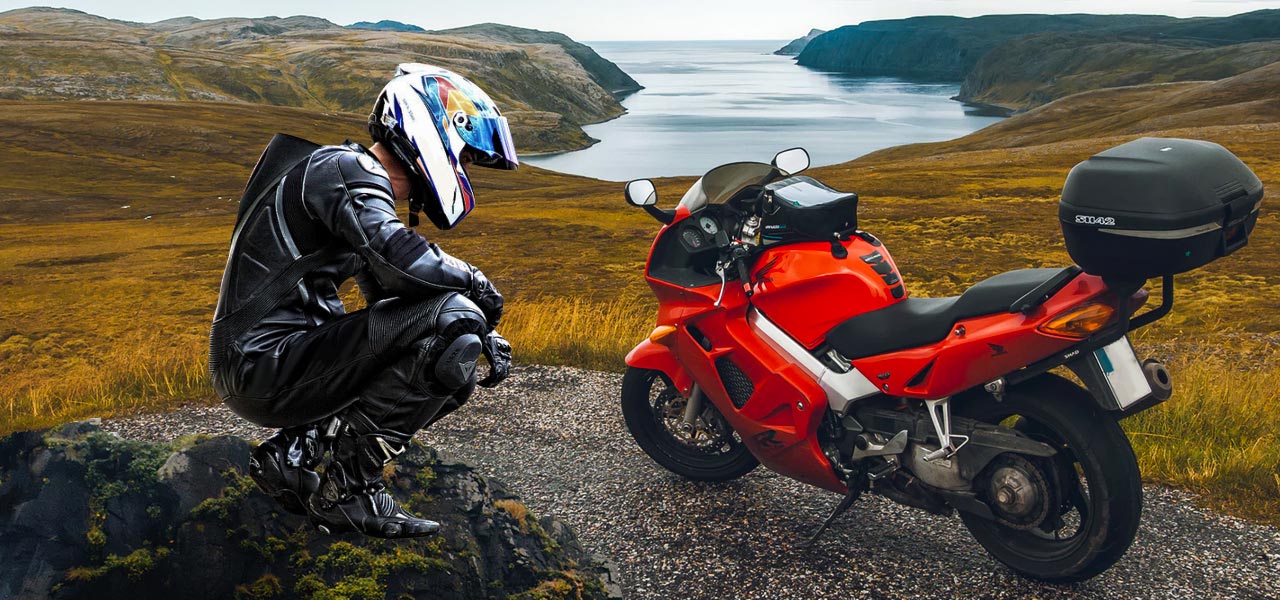 Un motorista de cuclillas junto a su moto en la montaña con todo el equipo ya que es un motero ATGATT